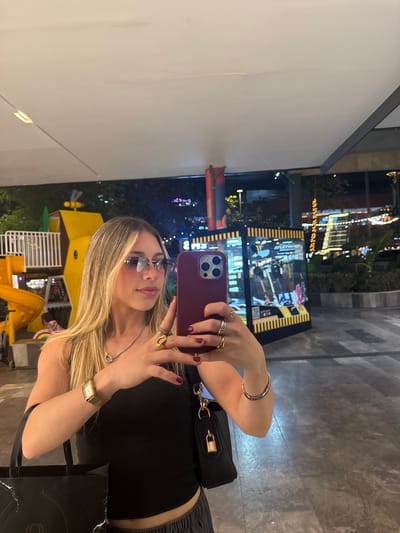 A young adult female with long, blonde hair and dark red fingernails is centered, taking a selfie with a maroon iPhone in her right hand. She wears a black ribbed tank top, grey pants, rectangular rimless sunglasses, a silver necklace with a circular pendant, multiple gold and silver rings, a gold-toned rectangular wrist watch, and a silver bracelet. She holds a black shopping bag in her left hand and a black handbag with a gold padlock accessory on her right side.

The background is an outdoor commercial or entertainment complex at night, illuminated by overhead lights and ambient city lights. To the left, a children's playground structure, predominantly yellow with white railings, a yellow slide, and a yellow bee-shaped ornament, is visible. A partially obscured figure is behind the playground. To the right, an illuminated display kiosk with black and yellow stripes showcases images of scooters or similar products. Further back, modern buildings with illuminated windows and decorative string lights are visible against the dark sky. The ground is paved with patterned grey tiles. This scene is situated within a modern commercial area, consistent with a shopping mall courtyard, in San Agustín, Mexico.