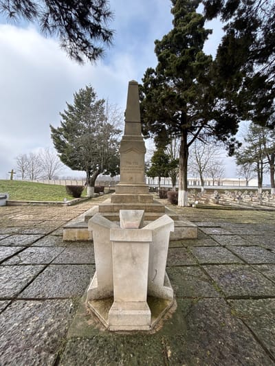 The image displays a war memorial situated within a cemetery in Shumentsi, Bulgaria, under an overcast sky. A central, weathered stone obelisk with a multi-tiered base features the inscription "1916-1918" and additional Cyrillic text on its main body. Directly in front of the obelisk is a lower, lighter-colored stone structure with four splayed supports. Both structures rest on a paved area composed of large, irregular dark grey stone slabs, showing moss accumulation in the crevices. To the right of the monument, numerous rows of small, uniform light-colored grave markers are visible across a grassy expanse, extending towards a white building with a flat roof in the distant background. To the left, a grassy slope rises, crowned by a white cross in the distance. The area is populated by various trees, including tall, dense evergreens and several bare-branched deciduous trees; some tree trunks exhibit white paint near their bases. Dark pine branches partially obscure the upper left corner of the frame. No people or active subjects are present in the scene.