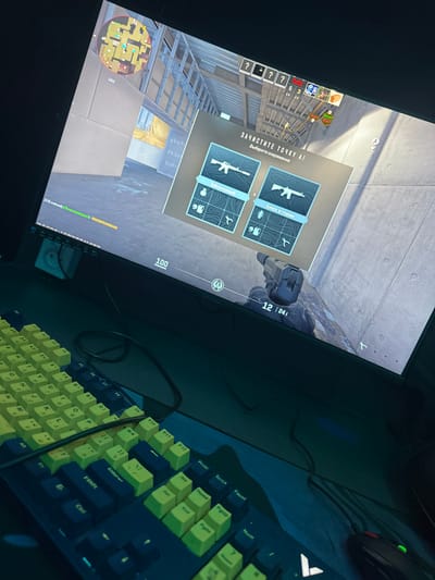 A slightly angled view shows a gaming setup. A computer monitor displays what appears to be a first-person shooter video game, showcasing in-game objectives and weapon choices. A keyboard with a dark blue frame and yellow keys is positioned to the right of the monitor. A mouse is also visible, partially obscured in the top right corner of the image. The scene is illuminated, emphasizing the vibrant colors of the keyboard and the details on the monitor.