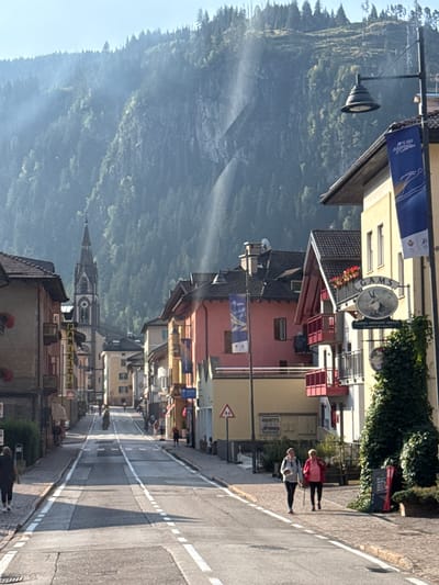 A sunlit street in the city of Predazzo, Italy, is lined with colorful buildings on either side. A prominent church spire rises in the background, partially obscured by trees. The street is paved and marked with white lines. Two women are walking on the sidewalk on the right side of the street. Several banners hang from lampposts, and a sign for "GAMS Pizzeria Ristorante Galleria" is visible on one building. The environment appears to be mountainous, with a heavily wooded slope forming the backdrop. The overall scene suggests a peaceful, daytime setting in a town nestled within a natural landscape.