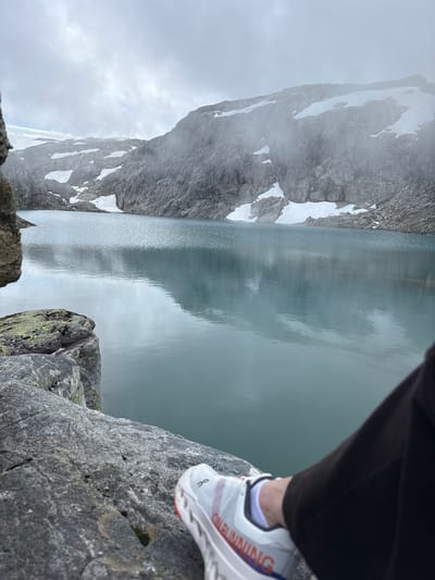 An outdoor, high-angle, medium shot captures a serene mountainous landscape with a person's shoe and leg in the foreground. The person is seated on a large rock on the left, their right leg extended towards the water. The shoe is white with orange and blue accents and the text "ON RUNNING." The environment features a calm, turquoise lake reflecting the overcast sky and mist-shrouded, gray, rocky mountains. Patches of snow are visible on the mountainsides, suggesting a high-altitude location. The overall impression is one of tranquil exploration in a remote, natural setting. The location is identified as Brattabø, Norway.
