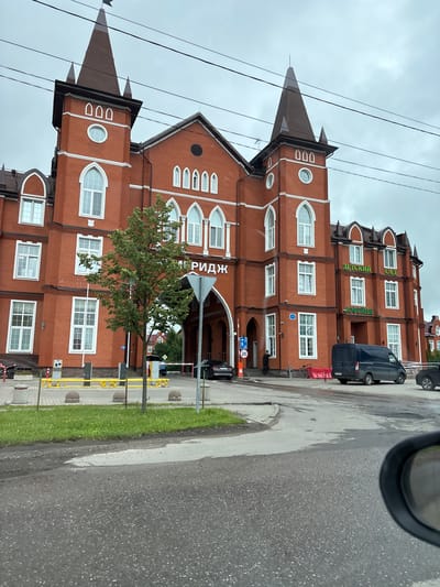 The image shows a red brick building with architectural elements resembling a castle, located in Krasnyi Posyolok, Russia. The building features multiple arched windows, decorative white trim, and conical towers topped with spires. Above a large archway leading into the building, the word "РИДЖ" is visible in large lettering. To the right of the archway, signs on the building read "ДЕТСКИЙ САД" (kindergarten) and "КОФЕЙНЯ" (coffee shop). A dark blue van is parked in front of the building, and a black car is visible in the background. A man is standing near the entrance to the building, and a bicycle is leaning against a red barrier. The ground in front of the building is paved, with a grassy area and a road in the foreground. The sky is overcast. The image appears to have been taken from a vehicle, as a side-view mirror is visible in the bottom right corner.