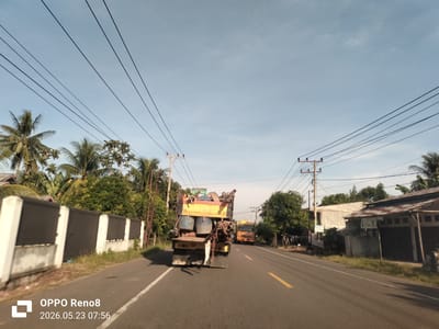 A medium-angle shot shows a road with yellow lines down the middle in Ierhobglumpang, Indonesia. A truck filled with pipes and other equipment is driving down the road, with another truck visible in front of it. Trees and buildings line either side of the road. Power lines are strung across the sky. The photo was taken with an OPPO Reno8, and the date stamp indicates the photo was taken on May 23, 2026, at 07:56.
