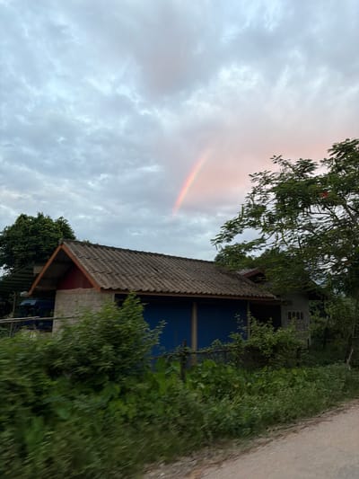 A faint rainbow arches across a cloudy sky above a rural scene in Hong Ngern Village, Laos. In the foreground, a simple, weathered house with a corrugated metal roof is partially obscured by lush green foliage. The house appears to be constructed of cinder blocks and wood, with blue shutters covering some of its openings. To the right of the house, a large, leafy tree with delicate branches extends into the frame. A dirt road runs along the bottom right of the image. The overall atmosphere is tranquil and somewhat overcast, with the subtle presence of the rainbow suggesting recent or impending rain.