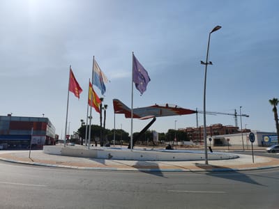 The image depicts a roundabout in Los Alcázares, Spain, featuring a prominent display of flags and a fighter jet monument. Four flags are visible: a red flag with a crest, a flag with blue and white stripes, the Spanish flag, and the European Union flag. The fighter jet, painted in red and gray, is mounted on a dark support structure in the center of the roundabout.
Surrounding the monument are palm trees and some shrubbery. Beyond the roundabout, there are buildings, including a modern-looking structure with a glass facade and a Lidl supermarket. Construction cranes are visible in the background, suggesting ongoing development. A lamppost with lights stands to the right of the frame. The sky is clear and blue. The road surface in the foreground is asphalt with visible cracks. The overall scene is characteristic of a public space in a Mediterranean coastal town.
