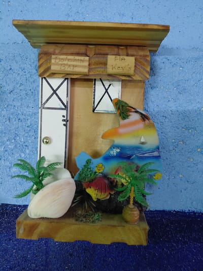 The image shows a souvenir diorama, likely depicting a scene from the Florida Keys, as evidenced by the "Fla Key's" inscription on a wooden plaque. The diorama is constructed from wood, with a backdrop showing a stylized depiction of a tropical island, including a dolphin jumping out of the water. The scene incorporates artificial elements such as miniature palm trees, a seashell, and a small fish. The base of the diorama is partially painted to resemble a dock or pier, with black X-shaped markings. The souvenir is resting on a textured, painted surface. There is no other contextual information to place the diorama within the city of Cutler Bay, United States of America.