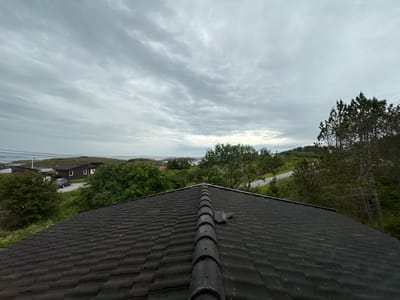 This is a medium-angle, eye-level, outdoor, wide shot of a dark, weathered rooftop. The rooftop is covered in dark grey shingles and has a ridge cap running along the peak. The ridge cap appears to be made of rounded, dark grey tiles, with one tile dislodged and tilted.

Beyond the rooftop, the scene unfolds into a rural landscape under a cloudy, overcast sky. The sky is filled with layers of grey clouds, suggesting a cool, possibly damp day.

In the middle ground, there are several low-lying buildings with dark roofs, similar in color to the foreground rooftop. A grey sedan is parked in the driveway of one of the buildings. Lush green trees and vegetation surround the buildings, with a winding paved road visible in the distance. The landscape appears to be coastal, with glimpses of water and islands in the far background. Electrical wires are visible strung across poles in the distance, indicating human infrastructure in the otherwise natural setting.

The overall impression is one of a quiet, slightly melancholic day in a coastal Norwegian village. There is no visible text in the image.