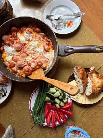 The image shows a breakfast spread on a wooden table. In a dark frying pan, there is a dish of scrambled eggs with sliced sausages and chopped tomatoes. A wooden spatula is placed in the pan. To the right of the pan, there is a plate with sliced bread and cheese. In the foreground, a plate holds chopped cucumbers, sliced tomatoes, and green onions. Another plate with a floral design and a fork is visible to the left. The environment is a domestic setting, possibly a kitchen or dining room, in Усть-Мана, Russia, as indicated by the food packaging in the lower right corner which contains Russian text. There are no people visible in the image.