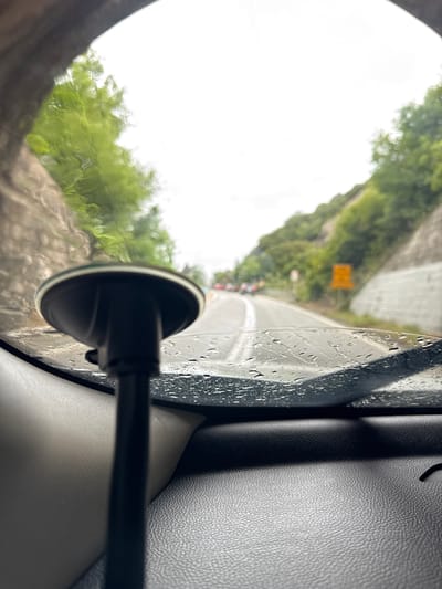 The image depicts a view from inside a car, likely a passenger seat, on a rainy day. Through the rain-streaked window, a road is visible, bordered by a rocky embankment with vegetation. A yellow road sign is partially visible in the distance. The interior of the car is visible, including the side window frame, a suction cup mount, and a portion of the interior door panel, possibly leather or a similar material. The location is presumably Prijevorac, Montenegro, based on the query.