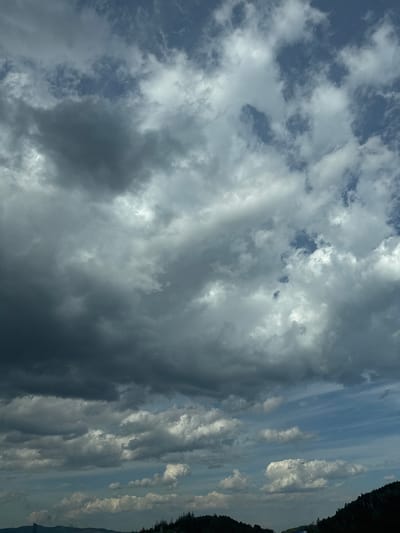 The image displays a cloudy sky over a forested hillside. Dark, heavy clouds dominate the upper portion of the frame, with lighter, scattered clouds appearing lower down. The lower part of the image shows the silhouette of a dark green forest against a hazy, distant landscape, suggesting a location in Dolno Varshilo, Bulgaria, characterized by natural topography. There are no people or specific objects readily identifiable within the scene.