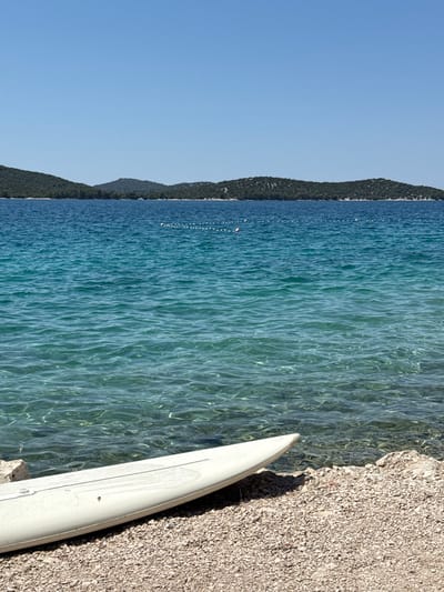 This is a landscape photograph taken on a sunny day in the city of Grad Šibenik, Croatia. The image features a clear blue sky dominating the upper portion of the frame, transitioning into the turquoise waters of the Adriatic Sea. In the foreground, a white surfboard lies partially on a rocky beach composed of small, light-colored pebbles. The surfboard is positioned diagonally, with its nose pointing towards the upper left of the image and its tail extending towards the bottom right. The water is exceptionally clear, allowing the pebbled seabed to be visible beneath the surface. Beyond the clear shallows, the sea deepens to a richer blue. In the distance, a chain of green, tree-covered islands forms the horizon line. A thin, dotted white line, likely a buoy line, stretches across the water between the foreground and the distant islands, with a few buoys visible. The overall scene evokes a sense of tranquility and a popular coastal destination.