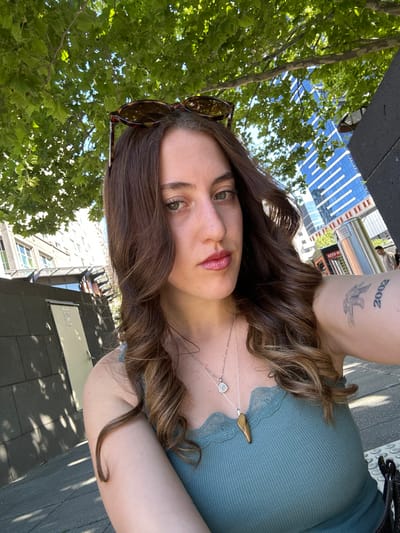 A woman with long, wavy brown hair and green eyes poses for a selfie. She wears a green top with lace trim and a silver necklace with a heart and leaf pendant. Sunglasses are perched on her head. A tattoo of "2002" and a bird is visible on her arm. In the background are trees with green leaves, a modern building with blue glass, and a street scene. The image is rotated counter-clockwise.