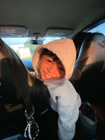 An overhead, close-up, wide-angle shot shows a young boy with a light-colored hooded sweatshirt in the passenger seat of a car. The boy is smiling and looking directly into the camera. The sun is setting, casting a warm glow on the boy's face. The interior of the car is visible, including the car's ceiling, a rearview mirror, and the back of the driver's seat. Outside the car, a blue sky with some trees and buildings is visible. The image was taken in Terban, Indonesia.