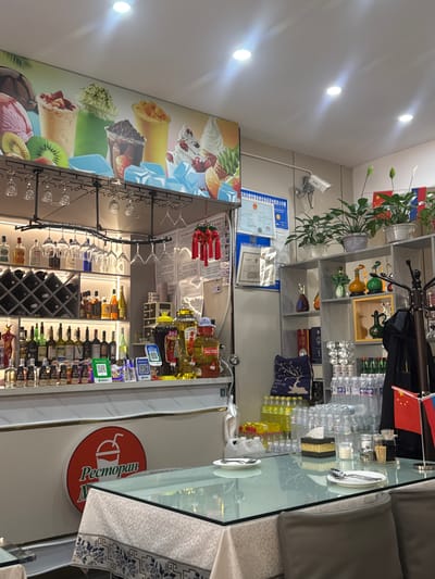 The image depicts the interior of a restaurant in Suifenhe, China. The scene is captured from a slightly elevated angle, looking across a glass-topped table towards a bar area and shelving units. On the table in the foreground, place settings with plates and cutlery are visible, along with a napkin dispenser and condiment shakers. The bar area is well-stocked with various bottles of alcohol, displayed on shelves and behind the counter. There are also several large decorative jars containing liquids and some smaller bottles. Above the bar, wine glasses are hung from a rack. The back wall features a large mural advertising various drinks and ice cream. To the right, shelving units hold decorative items, plants, and more bottles. A security camera is mounted on the wall above the mural. In the background, there are some flags, including the Chinese flag. The restaurant appears to be open for business, with food and drinks laid out on the table and bar.