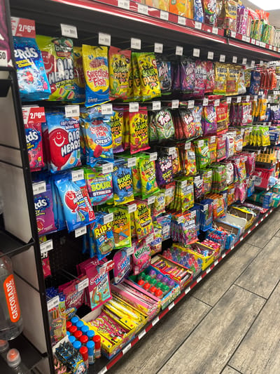 The image shows a shelf stocked with various candy products in what appears to be a convenience store or gas station. The items are displayed on black shelves with red trim. Among the visible candy brands are Airheads, Swedish Fish, Sour Patch Kids, and various gummy candies. Some of the products are displayed in clear plastic bags, while others are in boxes. Pricing tags are visible above some of the items. In the lower left corner, a partially visible bottle of Gatorade is present. The floor is tiled with a wood-grain pattern. The lighting suggests an indoor environment. The location is identified as Snyderville, United States of America. There are no people visible in the image.