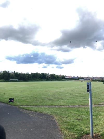 The image depicts an outdoor sports field, likely a football pitch, under a cloudy sky. The field is a large expanse of green grass with white goalposts visible in the mid-ground. There are several small white objects scattered across the field, possibly markers or equipment. In the background, a line of trees and houses are visible, suggesting an urban or suburban setting. Two birds are flying in the sky. In the foreground, a paved path curves along the edge of the field, and a gray metal post stands prominently. A black bag with a logo is situated near the path. A few individuals are present on the field, appearing to be engaged in some form of activity, though their actions are not clearly discernible from this distance. The overall scene suggests a typical day at a local park or recreational area. While the image is not explicitly geotagged to Shotton, United Kingdom, the presence of terraced housing and well-maintained sports fields is consistent with many locations in the UK.