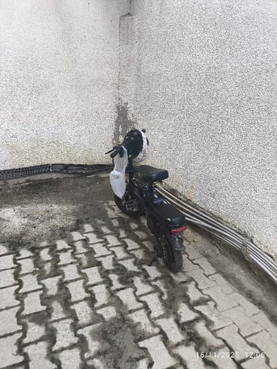 An electric moped, predominantly black, is parked facing diagonally left in the corner of an exterior space in Gonyeli, Northern Cyprus / Cyprus. A white helmet with dark markings is resting on the handlebars, from which a clear or translucent white plastic bag is suspended. The moped is supported by a kickstand.

The environment consists of two textured, light-colored stucco walls meeting at a right angle. The wall on the left features a run of black bundled cables secured horizontally along its lower section. The wall on the right shows several light-colored (likely grey or white) conduits running horizontally near the ground. Both walls exhibit vertical streaks of dirt or discoloration, particularly at the corner and lower sections.

The ground is paved with light grey, 'S'-shaped interlocking pavers. Darker, wet patches are visible across the pavers, indicating the presence of moisture. Distinct shadows of the moped and other objects are cast onto the ground and the lower part of the left wall, suggesting direct sunlight. No individuals are present in the scene. A timestamp of "16/11/2025 12:06" is visible in the lower right corner.