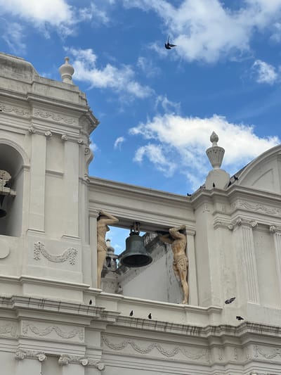 The image shows the exterior of the León Cathedral in León, Nicaragua. The cathedral is a large, white colonial-era building with ornate architectural details. Two statues of men are visible, supporting a large bell between them. Several birds are perched on the roofline and one is in flight in the blue sky with white clouds. The lighting suggests it is daytime.