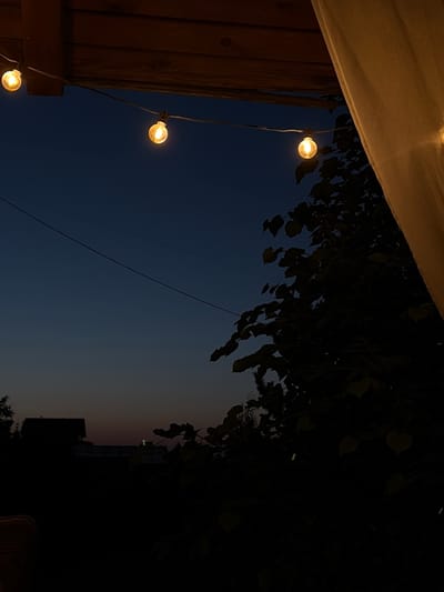 The image depicts an outdoor scene at dusk, likely in Leninsk-Kuznetskiy, Russia. String lights with warm, glowing bulbs are strung across the top of the frame, suspended from a wooden structure. To the right, a sheer, light-colored curtain is visible, partially obscuring the view. The background is a deep blue sky transitioning to a lighter hue near the horizon, indicating the last light of day. Silhouetted against the sky are dark masses of foliage from trees and bushes. In the distance, the dark shapes of buildings and possibly a roofline can be discerned. The overall atmosphere is calm and serene, suggesting an evening spent outdoors.