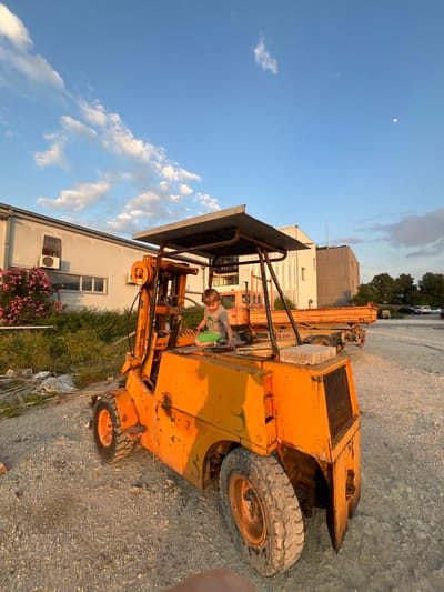 The image captures a young boy sitting in the driver's seat of an old, orange forklift parked on a gravelly lot in Pobrdje, Montenegro. The forklift is weathered and shows signs of rust. A building with a white facade and visible air conditioning units stands behind the forklift, accompanied by a bush with pink flowers. Beyond the immediate foreground, a taller building and some parked cars are visible, suggesting an urban or industrial setting. The sky is partially clear with scattered clouds and a faint moon visible in the upper left corner. The lighting suggests it is late afternoon or early evening. The boy is wearing a t-shirt and shorts, and he appears to be engaged in the act of playing or pretending to operate the machinery.
