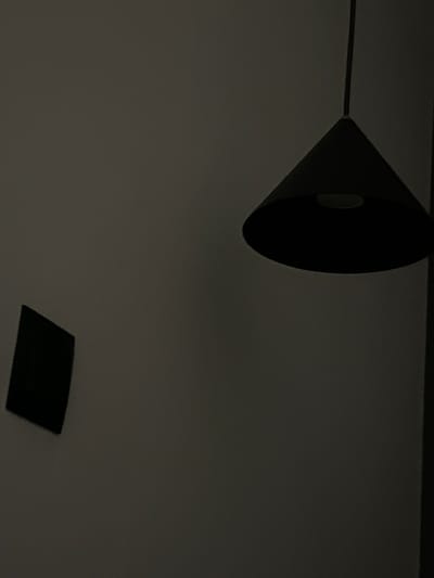 A dark, cone-shaped pendant light hangs from a cord against a gray wall. To the left of the light, a small, dark, rectangular object is affixed to the wall at an angle. The lighting is dim, suggesting an indoor environment with minimal ambient light. There are no people or animals visible in the image. The scene does not provide enough information to determine its location within the city of Siedlce, Poland.