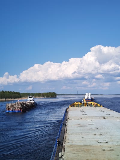 The image shows a wide river with two barges on it under a partially cloudy sky. One barge in the distance, carrying a load of logs, is headed away from the viewer. In the foreground is the deck of a larger, flat-topped barge with yellow and gray panels. The river flows through a green, forested area. The location is identified as Popovo, Russia.