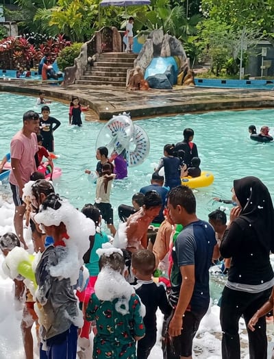 The photograph shows a crowded scene at a water park in Durin Simbelang, Indonesia. Numerous people, including children and adults, are in a pool filled with light blue water. Some individuals are swimming or wading, while others are standing on the pool's edge. Many people are covered in white foam. In the background, there is a structure with steps leading up to a platform, resembling a small waterfall feature. The environment includes lush greenery, with trees and plants surrounding the pool area. The weather appears sunny and bright. Several inflatable toys are visible in the pool.