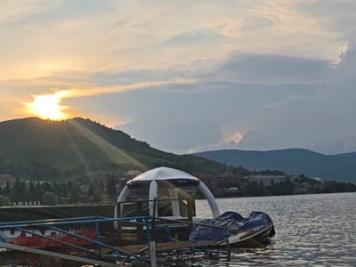 This is a medium shot, outdoor, horizontal photograph of a lake and mountainous terrain at sunset. The sun is setting behind a mountain on the left side of the frame, casting a warm glow across the sky. The sky is filled with clouds, some of which are illuminated by the setting sun. In the foreground, there is a pier with a white inflatable structure and two jet skis. The water of the lake is calm and reflects the colors of the sky. The mountainous terrain in the background is covered with trees and some buildings. The scene appears to be in the city of Якты-Куль, Russia.