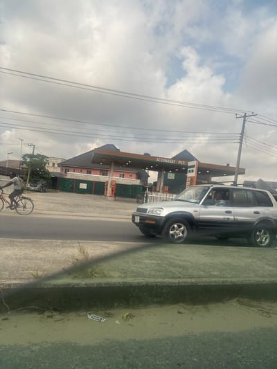 The image captures an urban street scene in Elimgbu, Nigeria, under an overcast sky. The viewpoint is from a low angle, likely from inside a vehicle, looking across a road towards buildings.

In the midground, an asphalt road features two visible lanes. A silver and black two-tone sport utility vehicle, consistent with a Toyota RAV4, occupies the lane closer to the viewer, traveling towards the left side of the frame. A male driver is visible through the windshield, along with a passenger seated to his right. In the far lane, a male cyclist rides a bicycle towards the right. He wears a light-colored short-sleeved shirt, dark trousers, and carries a dark bag slung over his left shoulder.

Beyond the road, a petrol station named "ACCONABAT PET." is prominent. It features a brown canopy supported by columns, sheltering at least two fuel pump islands equipped with orange and white dispensers. A green building is situated directly behind the pumps, and a two-story pink building is visible further back behind the station premises. Additional structures and trees are discernible in the background. Overhead, a network of utility poles and power lines spans the scene.

The immediate foreground reveals a concrete curb and a strip of dirt interspersed with sparse vegetation, where a white and blue wrapper is among scattered debris. A faint, elongated reflection is present in the lower portion of the image.