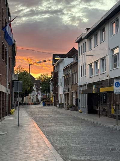 The image depicts a street scene in Sittard, Netherlands, during what appears to be sunset. The sky is filled with dramatic, colorful clouds, ranging from pale gray to vibrant orange and pink, suggesting either a beautiful sunset or the aftermath of a storm.
The perspective is from a low angle, looking up towards buildings that line a paved street. The buildings on the left are modern, with numerous windows and a white facade. On the right, the buildings are older, constructed of brick. The street itself is paved with cobblestones, and a line of warm light from the setting sun reflects on the pavement.
Several objects are visible: a Dutch flag is prominently displayed on the left, attached to a building. A construction crane is visible in the middle distance, rising above the rooftops and silhouetted against the colorful sky, indicating ongoing development or maintenance work in the area. Streetlights and various signs are present along the street. Some of the signs have text, but it is not clearly legible in the provided image. One sign visible at the top of the image appears to be a traffic sign with a pedestrian and bicycle symbol.
There are no people clearly visible in the frame. The overall mood is somewhat serene, with the dramatic sky creating a visually striking contrast with the urban environment. The presence of the crane suggests a functional, working town rather than a purely residential area. The time of day is clearly indicated by the sunset colors in the sky and the warm light on the street.