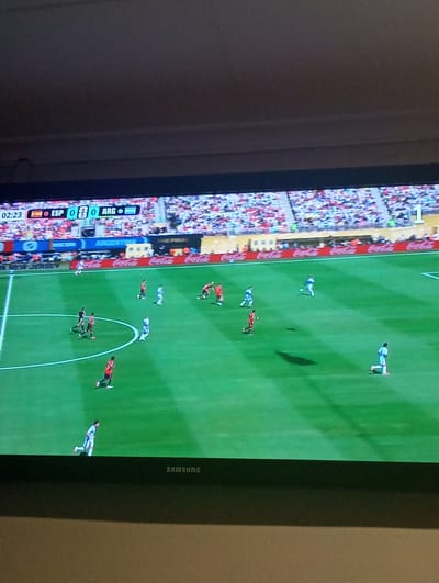 The image shows a soccer match on a television screen. The score displayed is ESP 0 - 0 ARG, with the time at 02:23. There are several soccer players on the field, some wearing red and others blue and white. The stadium is filled with spectators. The scene is indoors, with the television screen mounted on a stand or wall, as indicated by the Samsung logo at the bottom of the screen. The image does not provide any information about the location of Priego De Córdoba, Spain.