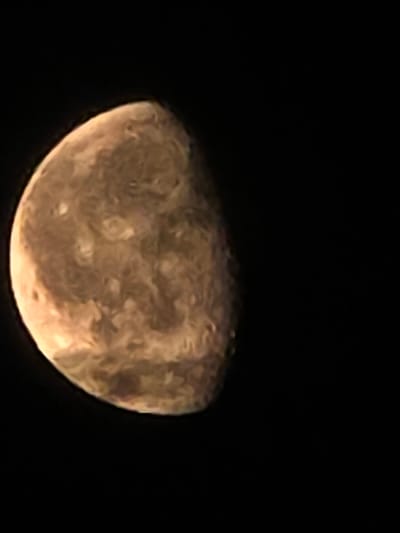 The image is a close-up of a celestial body, possibly a planet or moon, against a black background. The object is illuminated from the left, with a crescent shape visible. The surface of the celestial body appears textured with variations in color, ranging from light tan to a deeper brown. There are no discernable features that would place this image within the city of Икское Устье, Russia. The image focuses solely on the astronomical object and its immediate dark surroundings.