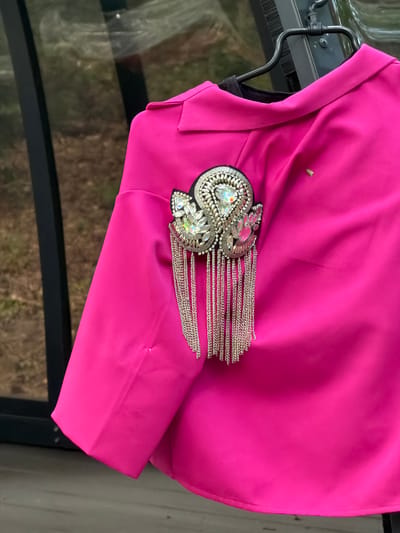 A bright pink jacket with a jeweled shoulder embellishment is displayed on a hanger. The jacket is made of a smooth, possibly satin-like material. The embellishment features a central large clear jewel, surrounded by smaller clear and pink jewels, and intricate silver beadwork. Silver chains hang from the bottom of the embellishment, creating a fringed effect. The jacket is hung on a black metal hanger, and the background suggests an outdoor or semi-outdoor setting with reflections in a glass surface, hinting at a location near a building or structure. The overall impression is one of a fashion item being showcased.