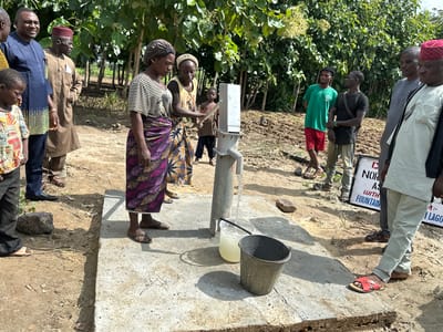 In Afogo, Nigeria, a group of people, including men, women, and children, gather around a newly installed water pump. The pump, made of metal, is situated on a concrete platform. A clear plastic bucket filled with water sits next to a metal bucket, both placed at the base of the pump. The scene is set outdoors, with lush green trees forming the backdrop. The ground is a mixture of dirt and sand, with some dry vegetation scattered around. The people are dressed in various traditional and casual attires, suggesting a community event or a demonstration of the new water source. The overall atmosphere appears to be one of shared interest and perhaps relief, as the water pump signifies a crucial resource for the community.