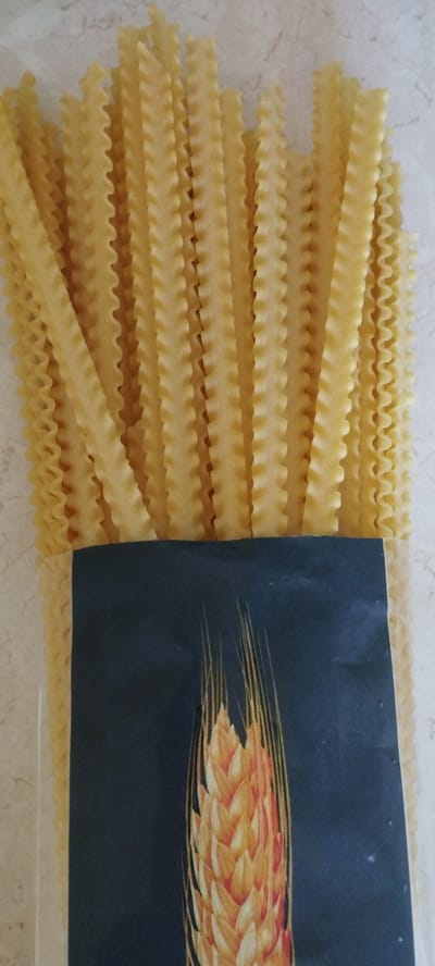 A package of dried pasta is displayed vertically, with the top portion showing the long, ruffled strands of pasta. The bottom portion of the package features a dark navy blue label with a stylized illustration of a wheat stalk in shades of yellow and orange. The pasta is light yellow and appears to be a type of mafaldine or similar ribbon pasta with wavy edges. The background is a light-colored, textured surface that resembles a countertop or table. The image is taken from a slightly angled overhead perspective.