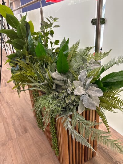 This is a media file of a decorative arrangement of artificial plants. The arrangement features a variety of green foliage, including large, glossy leaves and delicate fern-like fronds. There are also lighter, silvery-gray leaves that add contrast. The plants are densely packed and appear to be arranged in a bouquet or centerpiece. The background shows a light-colored, reflective surface, possibly a wall or a large piece of furniture, with a metallic railing. To the right, there is a wooden surface with horizontal slatted detail, and below that, a wooden floor. The lighting suggests an indoor setting, likely during the day. There is no visible text, people, or any specific activity or event taking place. The setting appears to be a domestic or commercial interior, possibly a lobby or a decorated space. The overall impression is one of artificial greenery used for decoration. The provided location context, Старое Мартьяново, Russia, does not offer any specific cues visible within the image itself.
