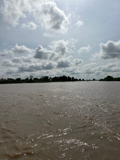 A wide, muddy river flows through Kunini, Nigeria, under a cloudy sky. The water has a choppy surface, reflecting the sunlight in shimmering patches. In the distance, a lush green treeline marks the far bank of the river, with a few buildings visible amongst the foliage on the left side of the image. The sky is filled with dramatic, cumulus clouds, some dark and heavy, others bright and white. The overall impression is one of a natural, rural landscape.