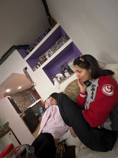 Two women are sitting on a couch in a dimly lit room. The woman on the right is wearing a red and gray jacket with a white emblem on the sleeve. She has long dark hair and is looking to the side with her hand on her chin. The woman on the left is wearing a light pink hoodie and black pants. She is leaning her head against her hand and appears to be asleep or resting. In the background, there are purple shelves on the wall filled with various items, possibly decorative objects or products. The room appears to be a living area or lounge.