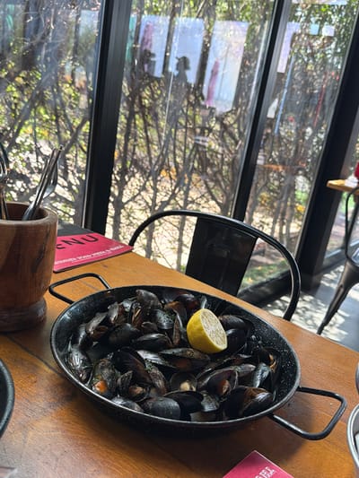 A close-up, eye-level shot shows a serving of cooked mussels in a black pan with handles, garnished with a slice of lemon. The pan sits on a wooden table, with a wooden utensil holder and a red menu to the left. In the background, through large glass windows, bare tree branches and foliage are visible, suggesting an outdoor or garden setting. The scene appears to be an outdoor seating area of a restaurant, likely in Umm Al Quwain, United Arab Emirates, given the natural light and vegetation.