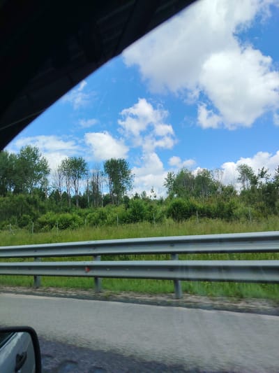 The image captures a view from a vehicle traveling along a road in Okulovka, Russia. In the foreground, a grey, metallic guardrail is visible, running horizontally across the frame. Beyond the guardrail, a grassy embankment slopes upwards, dotted with green bushes and tall trees. The trees are varied in density and height, with some appearing fully leafed and others exhibiting a more sparse, skeletal appearance. A section of the road surface is also visible, appearing to be asphalt. The sky above is a vibrant blue, interspersed with fluffy white clouds. The overall scene depicts a roadside landscape during daylight.