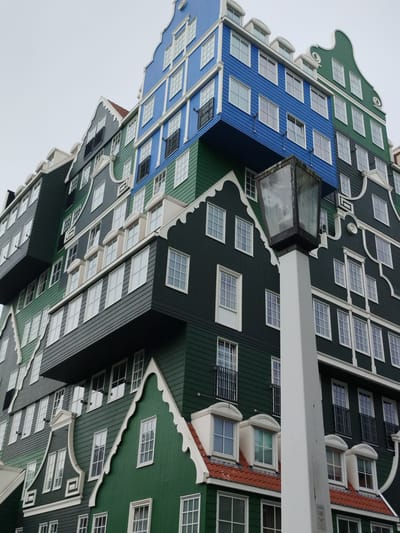The image shows a close-up, low-angle view of the Inntel Hotel in Zaandam, Netherlands. The hotel's distinctive architecture, designed to resemble a stack of traditional Dutch houses, is the primary focus. The building features multiple levels, each painted in different shades of green and blue, with white trim accenting the windows and gables. Numerous windows, varying in size and style, are visible across the facade. Small black balconies protrude from some of the windows. A tall, white lamp post with a glass lantern is positioned in the foreground, partially obscuring the building. The sky above is overcast and grey. The image is a static depiction of the building's exterior.