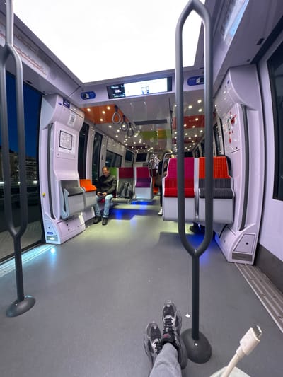 This image, rotated 90 degrees clockwise, depicts the interior of a modern train or tram carriage, likely within the public transport system of Nogent-sur-Marne, France. The carriage has a clean, contemporary design with light gray walls, ceiling, and floor, accented by integrated lighting.
Prominently in the foreground are dark gray, curved handrails and vertical support poles for standing passengers. The floor is a uniform gray. Seating consists of modular units with cushions in a vibrant palette of orange, gray, pink, red, and purple, arranged in various configurations, including face-to-face.
One male passenger is visible, seated on an orange and gray section, wearing a dark jacket and blue jeans, engrossed in his smartphone. In the immediate foreground, the feet of another person (likely the photographer) are visible, wearing gray sneakers, holding a white USB-C charging cable. A small blue panel on the seat structure features a charging symbol, indicating the availability of charging ports.
Digital display screens are integrated into the overhead panels, displaying information that is mostly unreadable. A schematic map, possibly of the train or metro lines, is affixed to a panel near the seating area. Small speaker grilles are visible on the ceiling. Large windows on the side of the carriage reflect a dark exterior, suggesting the scene takes place at night or in a tunnel. The overall environment appears modern, clean, and well-lit.