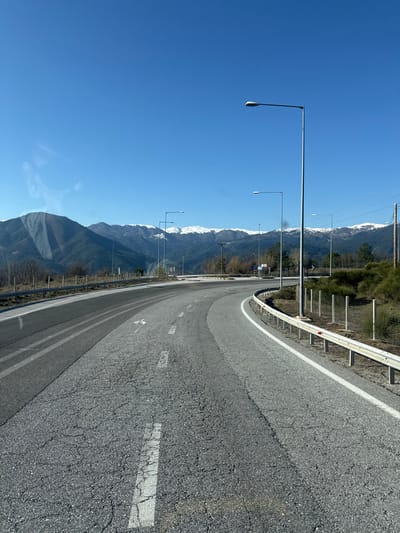 The image depicts a clear, sunny day on a winding asphalt road located in a mountainous area of Παναγία, Greece. The scene is observed from within a vehicle, indicated by faint reflections visible in the upper left quadrant. The road surface exhibits cracks and patches, and it curves distinctly to the right. A solid white line delineates the right shoulder, which is bordered by a metal guardrail supported by white posts. White dashed lines mark the center of the visible lane. Several tall, grey streetlights with horizontal arms and flat luminaires are regularly spaced along the right side of the road, extending into the background. Beyond the road, scrubby vegetation and sparse trees cover the foothills, leading to prominent mountains in the distance, some featuring snow-capped peaks under a bright, cloudless blue sky. Small blue road signs and wooden utility poles with overhead lines are discernible further along the road. No people or vehicles are present in the depicted scene.
