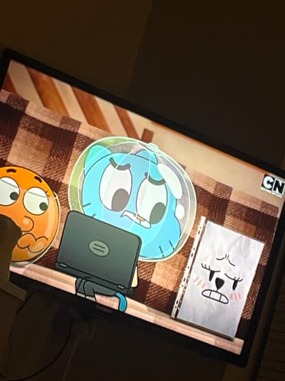 The image shows a television screen displaying an animated scene from "The Amazing World of Gumball." On the screen, two cartoon characters are visible: a blue cat-like creature with large eyes, enclosed in a clear bubble, and an orange, round creature with glasses, looking surprised. Between them is a laptop. To the right, a piece of crumpled paper with a drawing of a distressed face is propped up. The background of the animated scene is a patterned blanket. The television itself is set against a dark, out-of-focus background, likely a room. There is no information in the image to suggest its location in Biaga, Bulgaria.