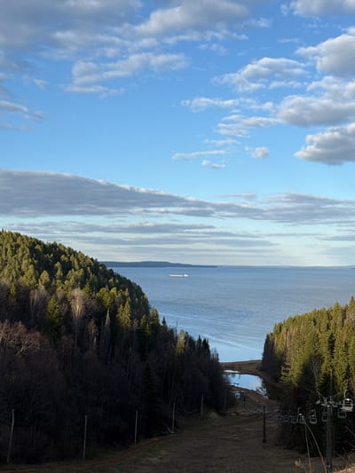 The image depicts a landscape from an elevated position, possibly a hill or mountain, overlooking a body of water. The sky is partly cloudy, with a mix of blue sky and white clouds. On either side of the water, there are forested areas with green trees. On the right side of the frame, there is a ski lift structure with chairs. The terrain on the right side appears to be a ski slope. A ship can be seen sailing at the horizon in the body of water. The image is taken in Galevo, Russia.