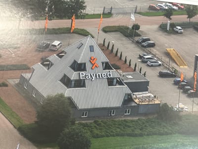 This is an aerial photograph of a modern, pyramid-shaped building with a grey, corrugated metal roof, located in Staphorst, Netherlands. The building has a distinctive orange logo on its facade, resembling a stylized figure with outstretched arms, and the word "Payned" is prominently displayed in large white lettering above it. Several windows are visible on the angled surfaces of the roof, and the lower section of the building is grey with white-framed windows.

The surrounding scene includes a paved parking lot with several cars parked, some of which are dark-colored SUVs and sedans, and a few smaller cars. An elevated wooden deck with picnic tables and benches is attached to one side of the building. Two orange flags on poles stand in front of the building. In the background, there are more parking areas, some trees, and a fence. The weather appears to be clear, and the time of day is likely daytime, given the lighting. The overall impression is of a commercial or business establishment in an industrial or semi-rural setting.