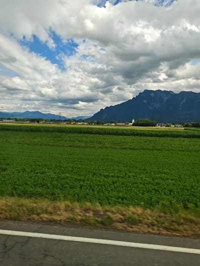 The image captures a panoramic view of a rural landscape under a cloudy sky, likely taken from a moving vehicle on a road. The foreground is dominated by a dark asphalt road with a white line, suggesting motion blur. Adjacent to the road is a strip of dry, brown grass. Beyond this, a vast expanse of vibrant green fields stretches out, possibly crops or pastures. In the mid-ground, a line of trees partially obscures a collection of buildings, including a white church with a steeple, indicative of a village or small town. Further in the distance, more fields and trees are visible, leading towards a range of imposing, dark mountains that form the backdrop of the scene. The sky is a dramatic mix of bright blue patches and heavy, grey clouds, hinting at changeable weather. The overall impression is one of a tranquil, agricultural area with a majestic mountain range.