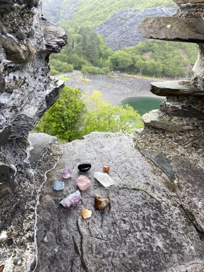 The image, taken from an elevated rocky outcrop in Llanberis, United Kingdom, overlooks a quarry landscape featuring trees, a quarry slope, and a body of water. A collection of small crystals and gemstones are arranged on the gray rock surface in the foreground. The rough, layered rock forms a natural frame for the broader landscape view.