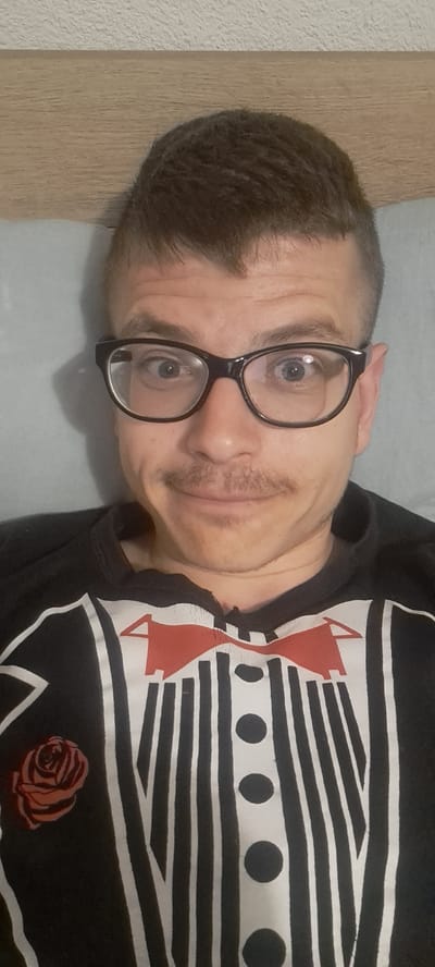 A man is seen lying down, wearing glasses and a black t-shirt with a white and red design. The design on the shirt includes stripes, circles, a bow tie, and a rose. The man has short, dark hair and a mustache. The location is not evident from the image, and there is no information to indicate Mineralni Bani, Bulgaria. The environment consists of a light-colored surface, possibly a pillow or bedding, and a wooden panel on the side.