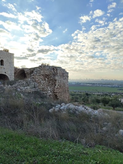 The image shows an old, partially ruined stone structure set against a backdrop of a cloudy blue sky. Part of a cityscape is visible in the distance. The structure appears to be crumbling, with missing stones and visible interior spaces. It is surrounded by grass and bushes. The image is rotated counter-clockwise.